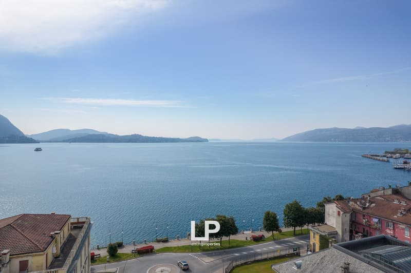 The image shows a scenic view of a lake surrounded by mountains. There is a small boat on the water and buildings along the shoreline. The sky is clear with some clouds. The landscape appears to be a picturesque lakeside town or village.