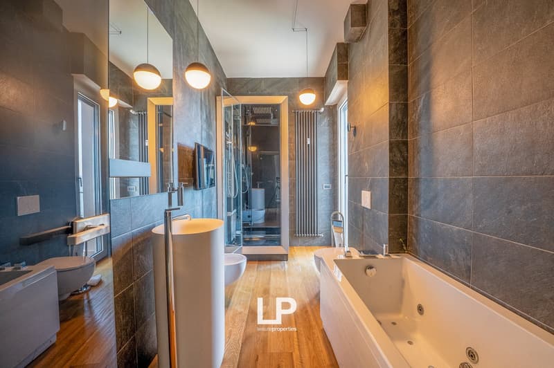 Spacious bathroom with a freestanding bathtub, glass-enclosed shower, vanity with sink, and tiled walls and floors. The room has modern lighting fixtures and a TV mounted on the wall.