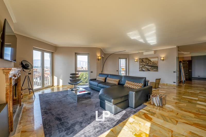 Spacious living room with large windows, hardwood floors, gray sectional sofa with patterned throw pillows, coffee table, and artwork on the walls. The room has a modern and stylish design with a balcony or terrace visible through the windows.