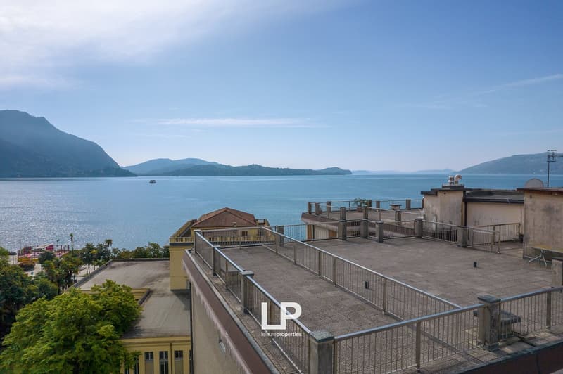 Multi-story building with balconies overlooking a scenic lake surrounded by mountains. The building has a rooftop terrace with railings, offering a panoramic view of the lake and the mountainous landscape.