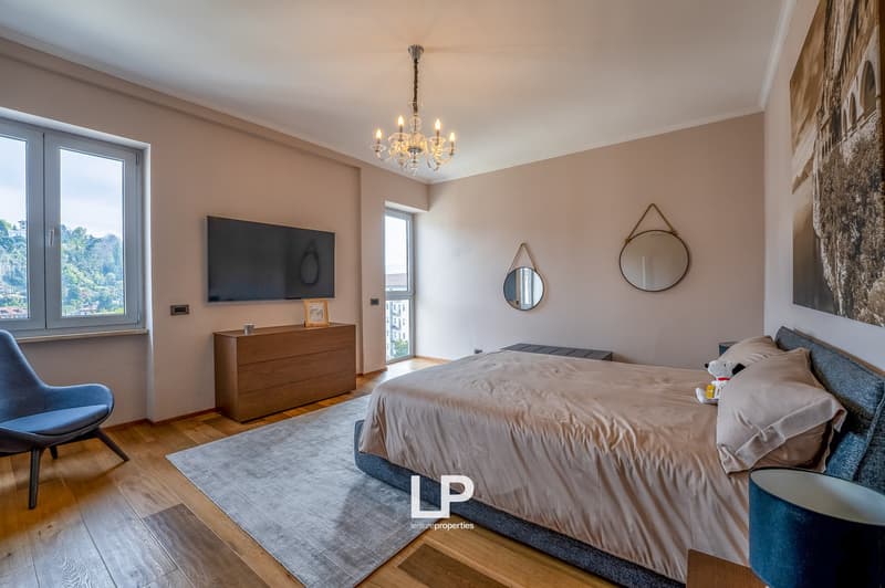 Spacious bedroom with hardwood floors, beige walls, large windows, a chandelier, a TV mounted on the wall, a dresser, and round mirrors on the wall. The room also has a balcony and a fireplace.