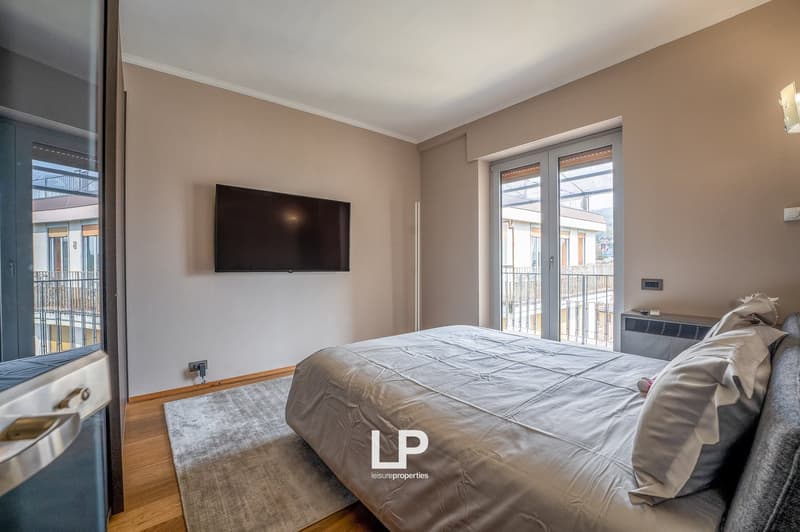 Spacious bedroom with large windows, hardwood floors, and a balcony. The room features a king-size bed, a mounted TV, and a cozy seating area.