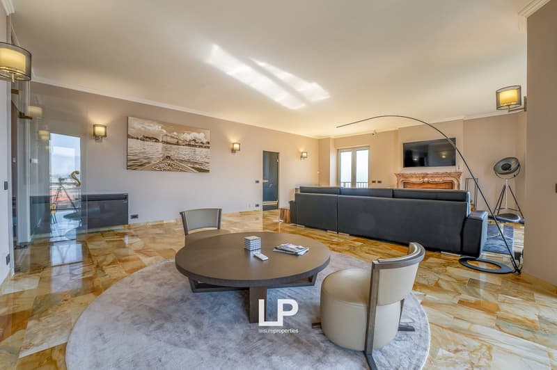 Spacious living room with large windows, hardwood floors, modern furniture including a round coffee table and comfortable sofas, and a fireplace. The room has a balcony or terrace with a view.
