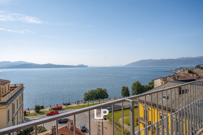 The image shows a scenic view of a lakeside town with mountains in the background. The apartment has a balcony or terrace that offers a panoramic view of the lake and surrounding landscape.