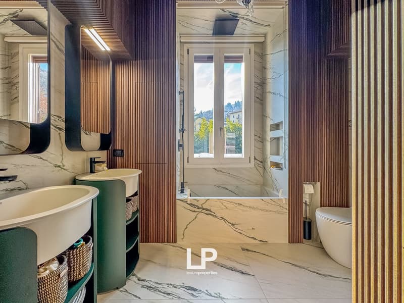 modern bathroom with two sinks, bathtub, toilet, large mirror, wooden wall panels, marble flooring, window with view