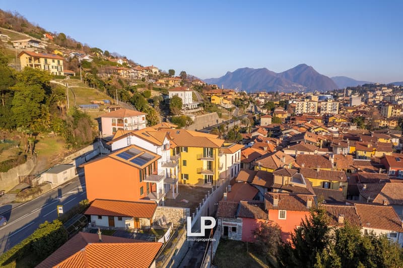 Hilltop terraced buildings with tiled roofs, some with solar panels, surrounded by greenery and distant mountain range