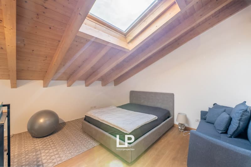a bedroom with a bed, skylight, gray couch, lamp, large exercise ball, and rug. hardwood floors and white walls. a wall power outlet and a nightstand outlet. a desk with an object on it and an open closet on the left side. a wooden ceiling with white wall