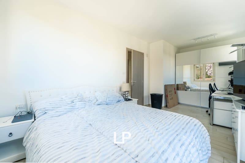 White walls, wooden floor, large bed, nightstands, a lamp, a trash bin, open door, and a small office area in the background.
