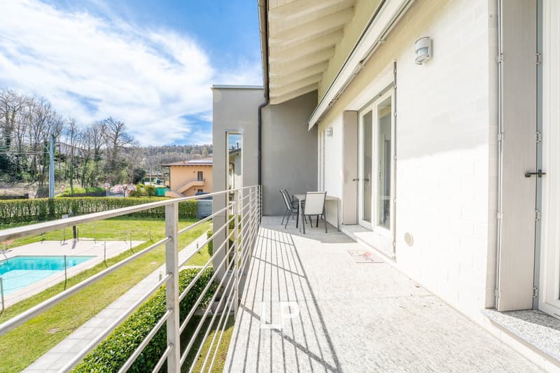 Balcony with metal railing, outdoor dining table and chairs, swimming pool, grassy area, trees, distant buildings
