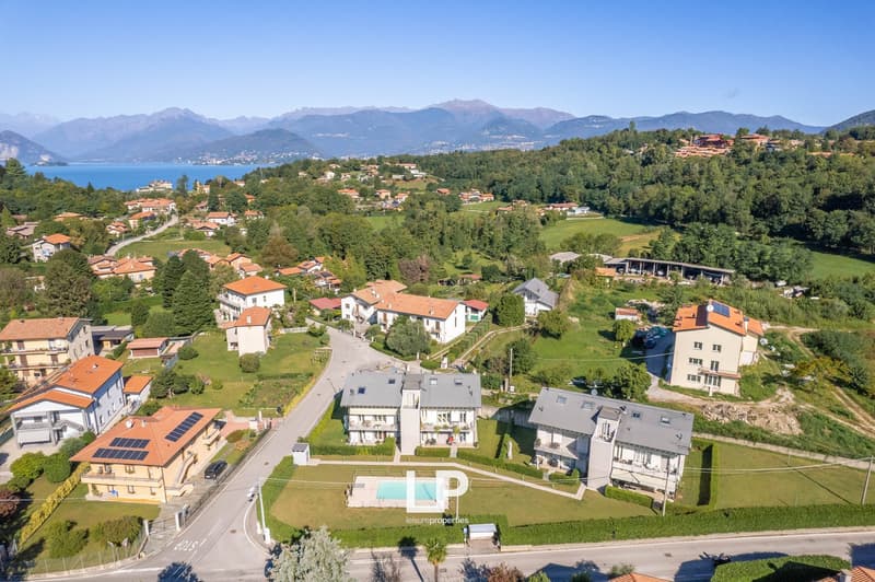 The image shows a scenic residential area nestled in a lush, mountainous landscape. The area features a mix of multi-story apartment buildings and individual houses with red-tiled roofs. There are paved roads, green lawns, and a few visible balconies or t