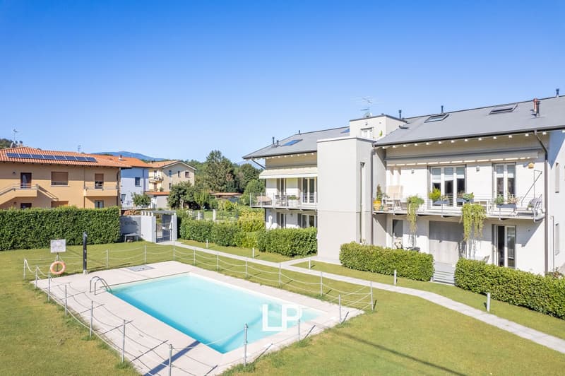 A multi-story apartment building with a swimming pool in the foreground surrounded by a well-manicured lawn and landscaping. The building has a modern, white exterior with balconies and large windows.