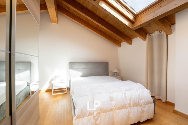 Bedroom with white walls, wooden floor, wooden beams, large window, skylight, a bed, two nightstands, and a lamp.