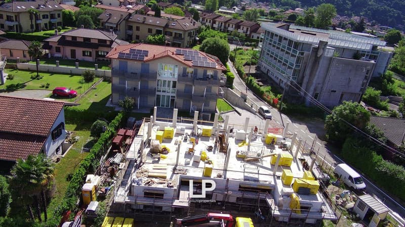 The image shows a construction site with various construction materials and equipment, including yellow containers and vehicles. The site is surrounded by residential buildings and a larger commercial or office building in the background.