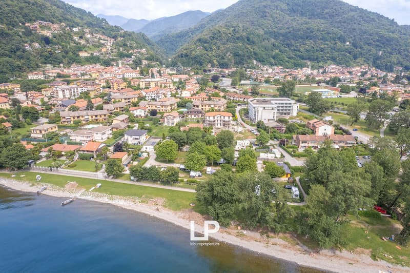 Aerial view of a mountainous town with densely packed residential buildings, hotels, and other structures. The town is situated along a lake or river, with a green park area visible. There are several multi-story buildings and apartments visible, as well