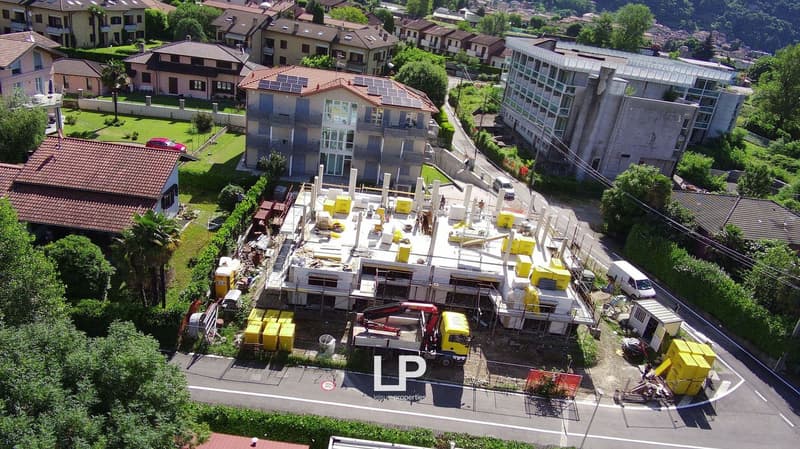 The image shows a construction site in a residential area. There are several buildings, some with solar panels on the roofs, surrounded by green spaces. The construction site is located on the ground level, with various construction equipment and material