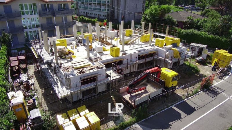 The image shows an active construction site with scaffolding, construction materials, and equipment such as cranes and trucks. There appears to be a parking area visible as well.