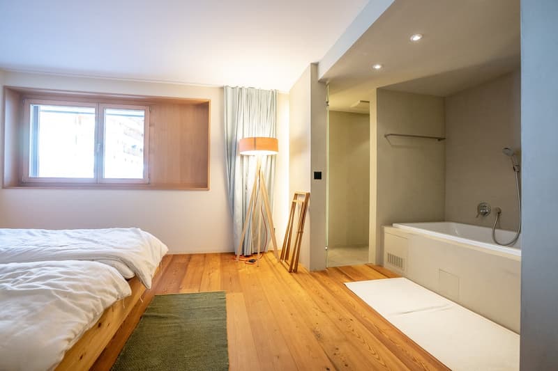 Spacious bedroom with hardwood floors, large windows, a bed, and an en-suite bathroom with a bathtub.