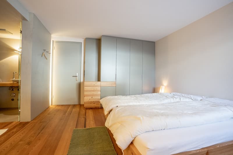 Spacious bedroom with hardwood flooring, gray built-in wardrobes, a large bed with white bedding, and a green rug on the floor. The room has a minimalist and modern design aesthetic.
