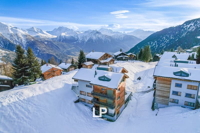Snow-covered mountain village with wooden chalets, apartment buildings, and scenic mountain views in the background