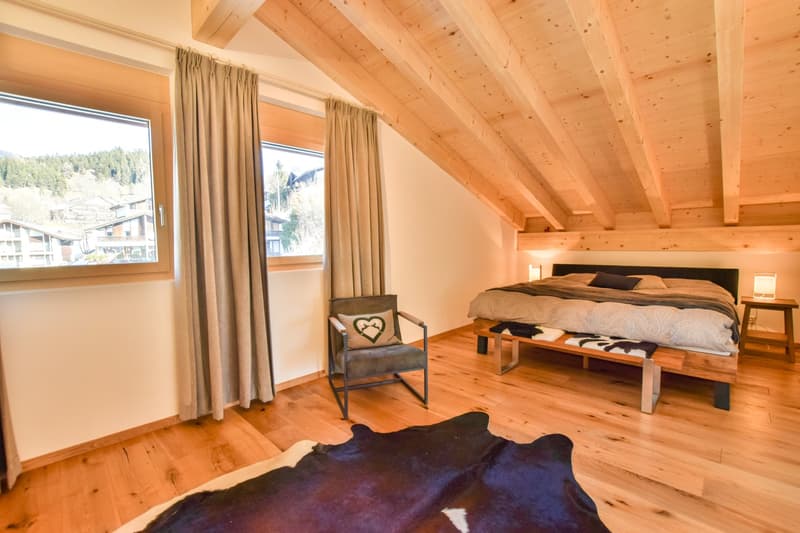 Spacious bedroom with wooden vaulted ceiling, large windows providing a scenic view, wooden floor, a king-size bed, and a cozy armchair.