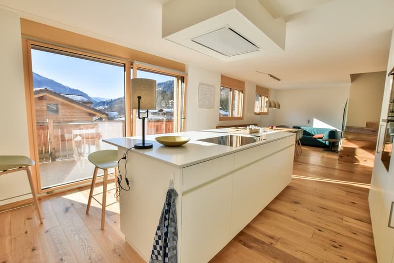Spacious open-plan kitchen with modern white cabinets, island counter, and large windows overlooking a balcony or terrace with a scenic mountain view.