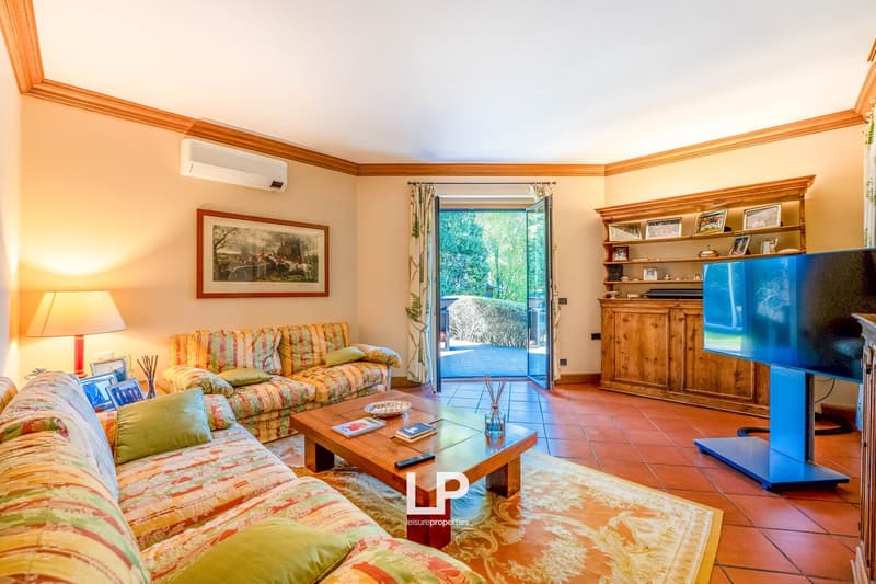 Spacious living room with large windows overlooking a balcony or terrace, colorful patterned sofas, wooden furniture, and a flat-screen TV mounted on the wall.