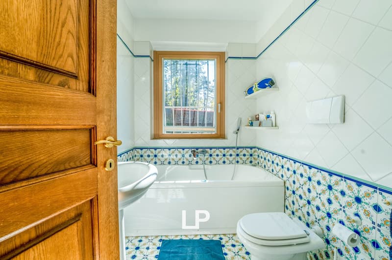 Bathroom with white tiles, wooden door, patterned tile border, toilet, and a window overlooking the forest outside