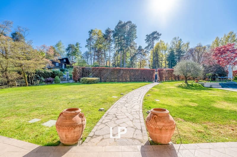 Large property with a stone path leading to a house surrounded by lush greenery, including trees, shrubs, and a well-manicured lawn. Two large terracotta pots are placed on either side of the path. The property appears to have a balcony or terrace and a p