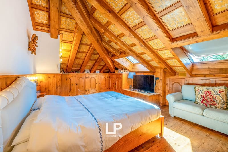 Spacious attic bedroom with wooden beamed ceiling, wooden cabinets and shelves, large bed, armchair, and a fireplace