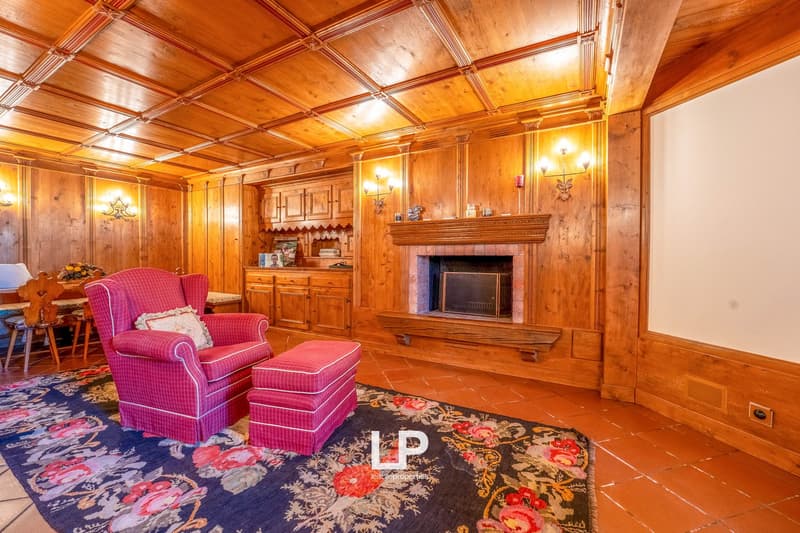 Spacious living room with wooden paneled walls and ceiling, ornate fireplace, built-in cabinets, and plush pink armchairs with matching ottoman on a patterned area rug