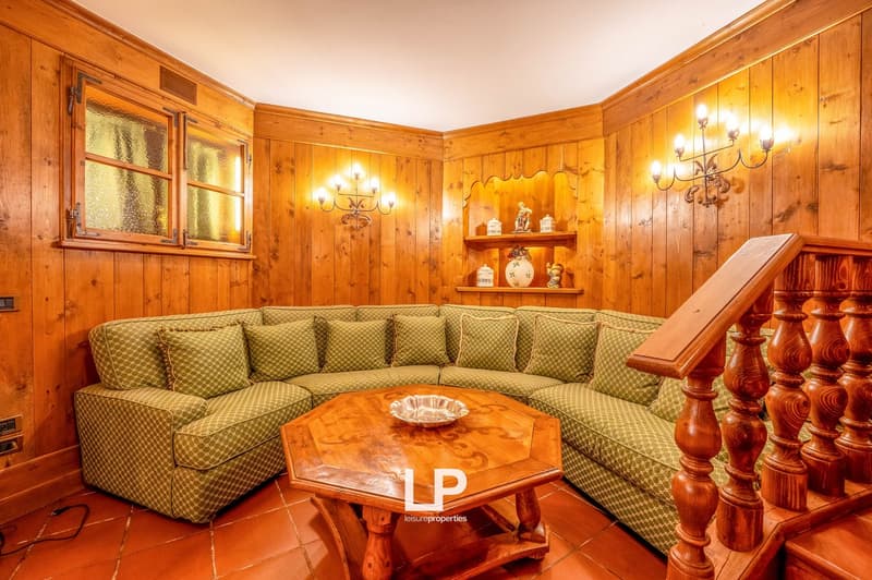 Cozy living room with wooden paneled walls, large sectional sofa with patterned upholstery, wooden coffee table, and decorative shelves with ornaments. The room features a fireplace and chandeliers, creating a warm and rustic ambiance.
