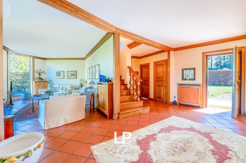 Spacious living room with high ceilings, wooden beams, and large windows overlooking the outdoor area. The room features a comfortable sofa, coffee table, and wooden furniture. The floor is covered with terracotta tiles, and there is a decorative rug in t