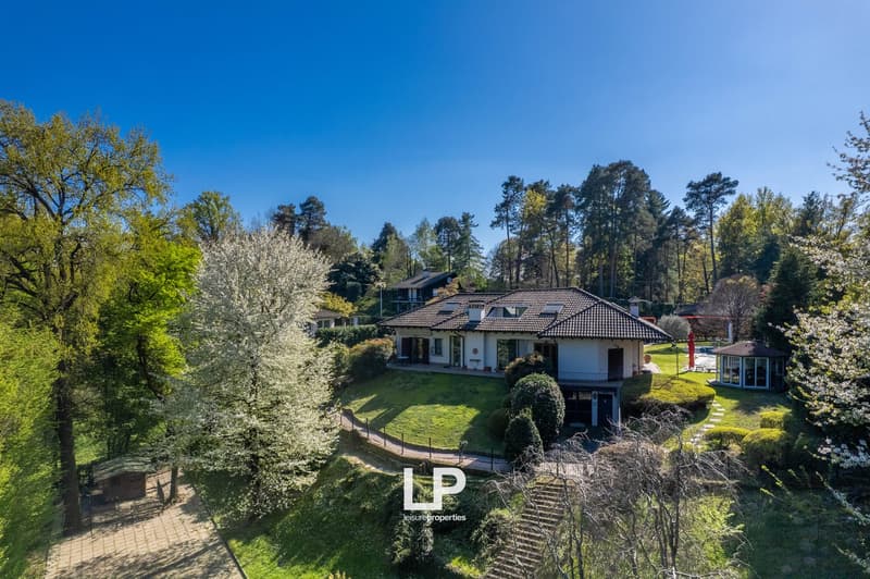Two-story house with tile roof, surrounded by lush greenery and blooming trees, with a paved walkway leading to the entrance and a parking area visible.