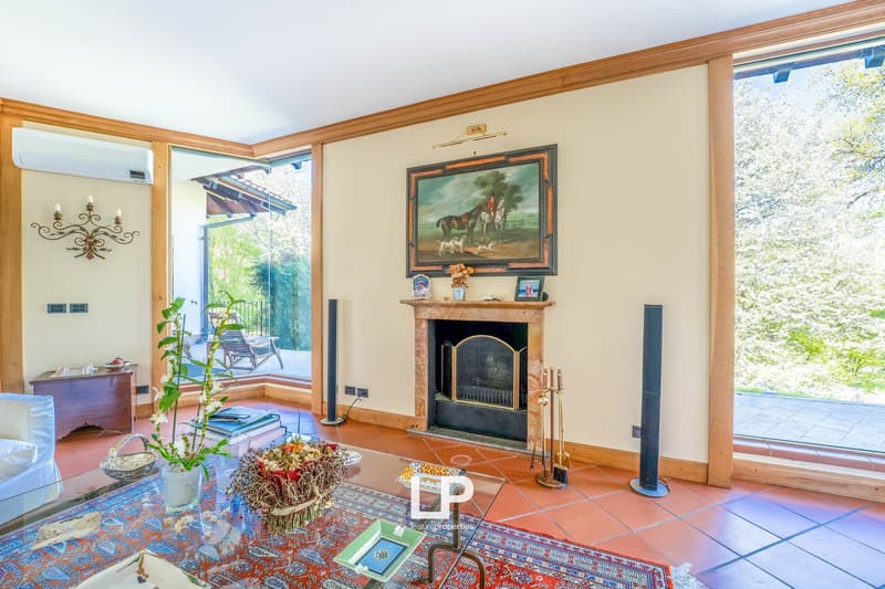 Spacious living room with wooden beams, large windows overlooking a balcony, a fireplace with a decorative painting above it, and a mix of traditional and modern furnishings.