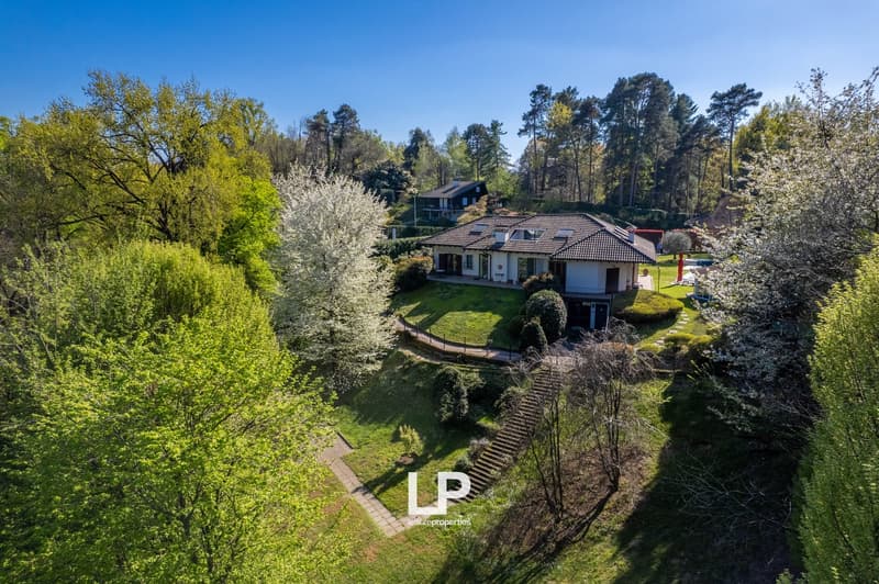 Two-story house with tile roof, surrounded by lush greenery and blooming trees, with a paved path leading to the house and a parking area visible.