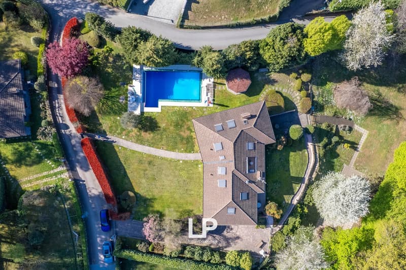 2-story house with a red-tiled roof, surrounded by lush greenery and landscaping. There is a swimming pool in the backyard and a driveway with parking spaces in the front.