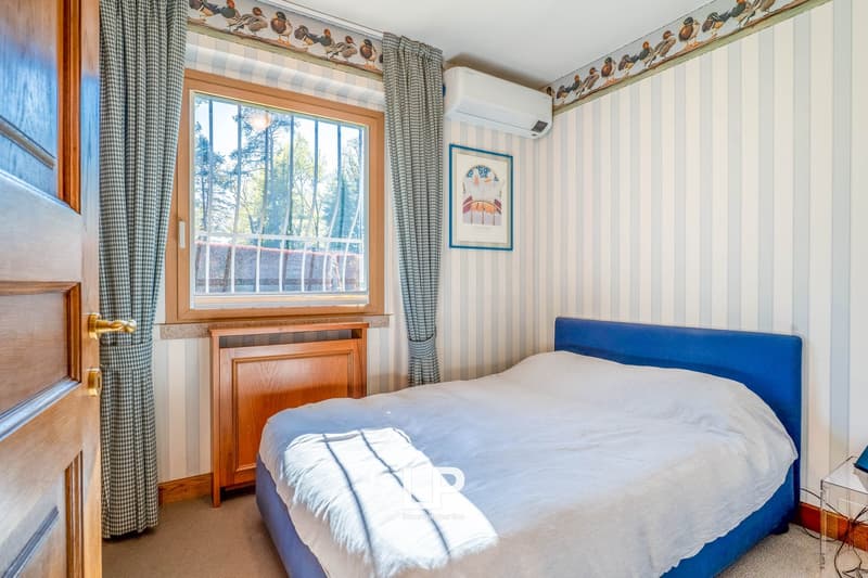 Bedroom with wooden furniture, blue upholstered bed, large window overlooking trees, patterned curtains, and wall decor