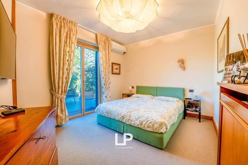 Spacious bedroom with a large bed, wooden furniture, and a sliding glass door leading to a balcony or terrace. The room has a warm, cozy atmosphere with beige walls and curtains.