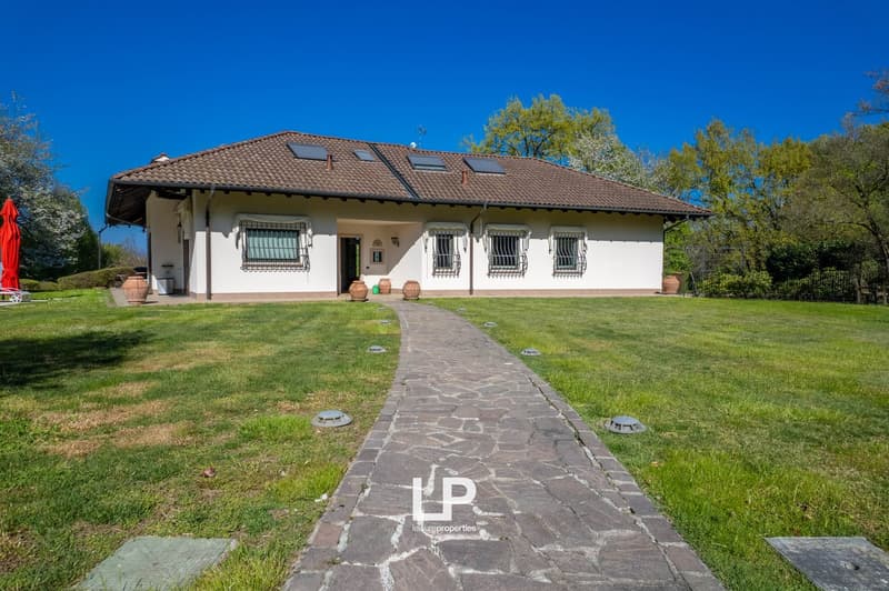 Single-story house with a tiled roof, white exterior walls, and several windows. There is a stone pathway leading to the entrance and a grassy lawn surrounding the house. The property also has a balcony or terrace visible.