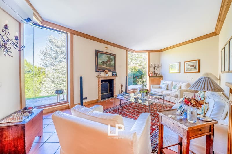 Spacious living room with large windows overlooking a garden, hardwood floors, white walls, wooden trim, fireplace, plush white sofas, glass coffee table, and various decorative elements such as artwork and lamps.