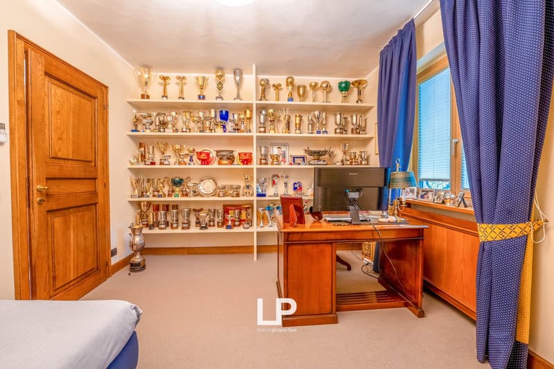 Spacious home office with wooden furniture, including a desk and shelves displaying numerous trophies and awards. The room has wooden doors, curtains, and a fireplace visible.