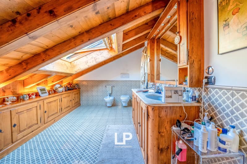 Spacious attic bathroom with wooden beamed ceiling, tiled floor, vanity unit, toilet, and various bathroom accessories and supplies visible.