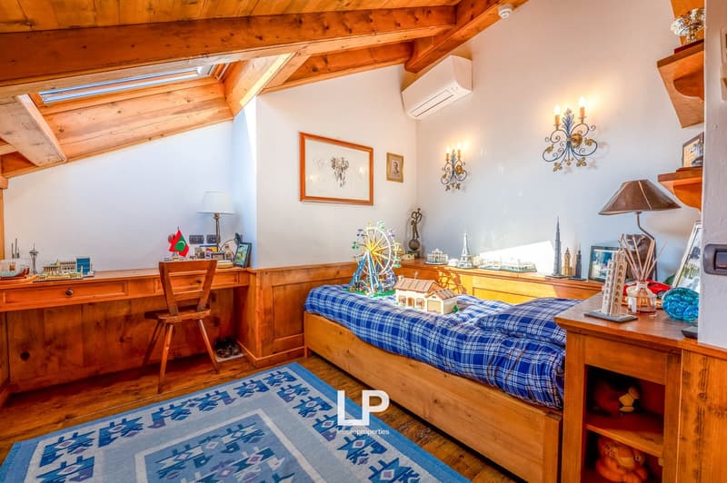 Cozy bedroom with wooden beamed ceiling, wooden furniture including a bed with plaid bedding, desk, and shelves displaying decorative items. The room also features a fireplace and wall decor.