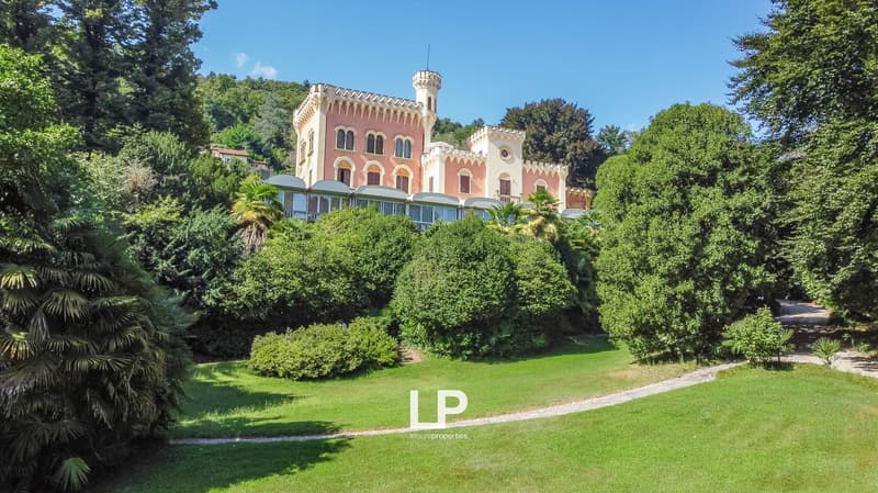 A large, ornate castle-like building with pink and white walls, towers, and turrets, surrounded by lush green trees and vegetation. The building appears to be situated on a hill or raised ground.