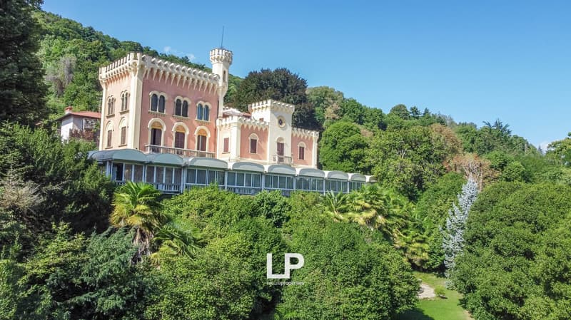A large, ornate castle-like building with towers and turrets, surrounded by lush greenery and palm trees. The building has multiple balconies and terraces.