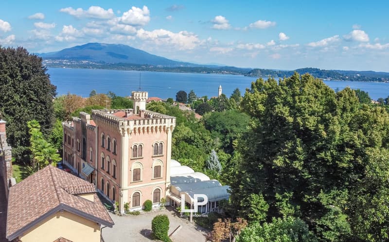 A large, historic castle-like building with red-tiled roofs and towers, surrounded by lush greenery and overlooking a scenic lake and mountains in the background.