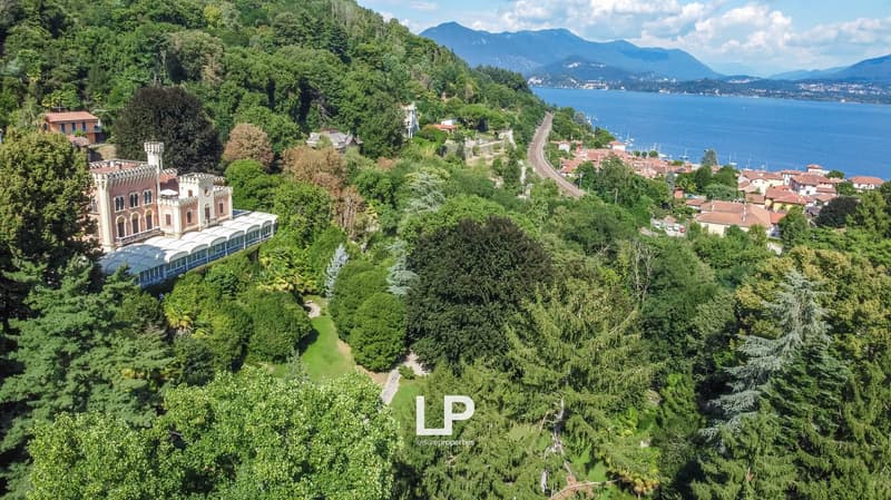 Aerial view of a scenic lakeside town surrounded by lush green forests and mountains. The town features a historic castle-like building and a winding road along the shoreline. The landscape is characterized by dense vegetation, including trees and shrubs,