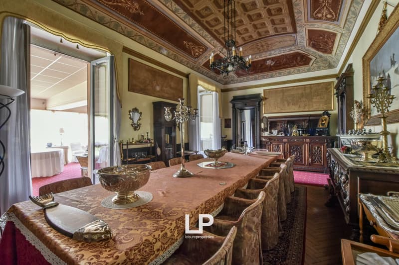 Ornate and luxurious dining room with intricate ceiling designs, antique furniture, chandeliers, and a large wooden dining table with chairs.