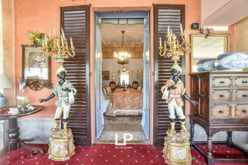 Ornate and lavishly decorated interior with wooden shutters, antique furniture, chandeliers, and decorative statues. The room features a large dining table with a patterned tablecloth and appears to be a formal dining or living space.