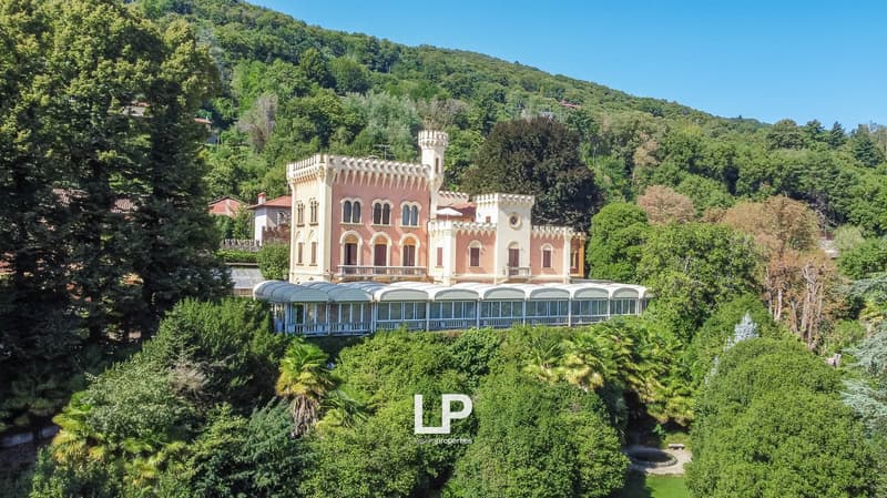 A large, ornate castle-like building with a pink exterior and white architectural details, surrounded by lush greenery and trees. The building has multiple floors, towers, and balconies, and appears to be situated in a scenic, mountainous area.